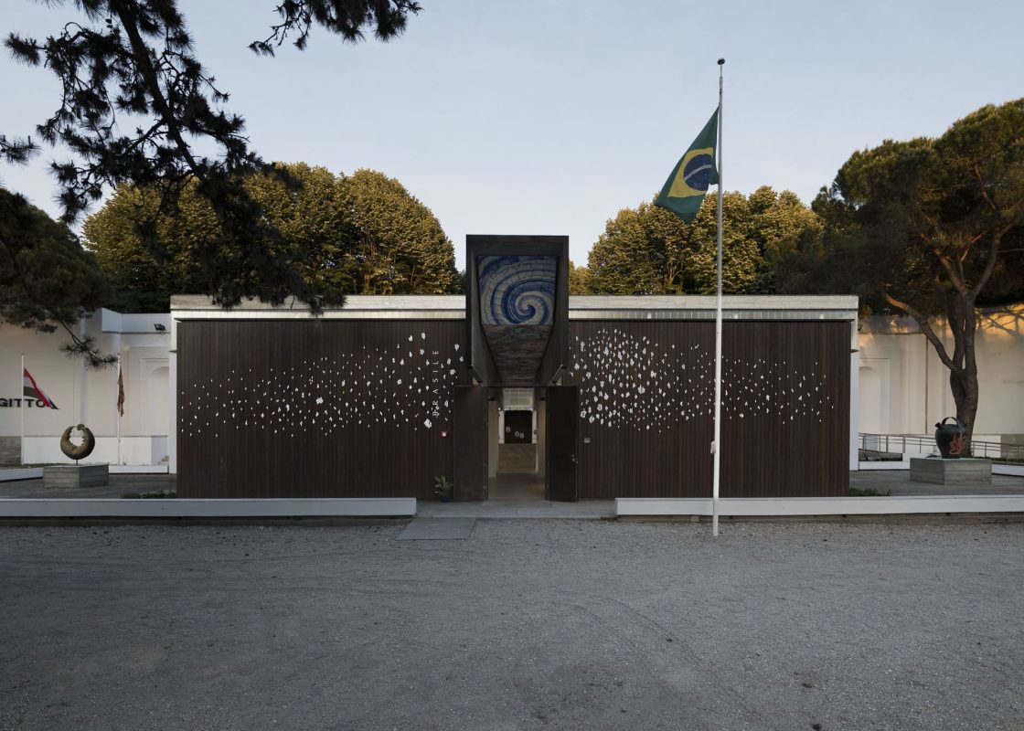 Vista externa do Pavilhão do Brasil na Bienal de Veneza 2026, com fachada modernista em madeira escura e bandeira brasileira hasteada