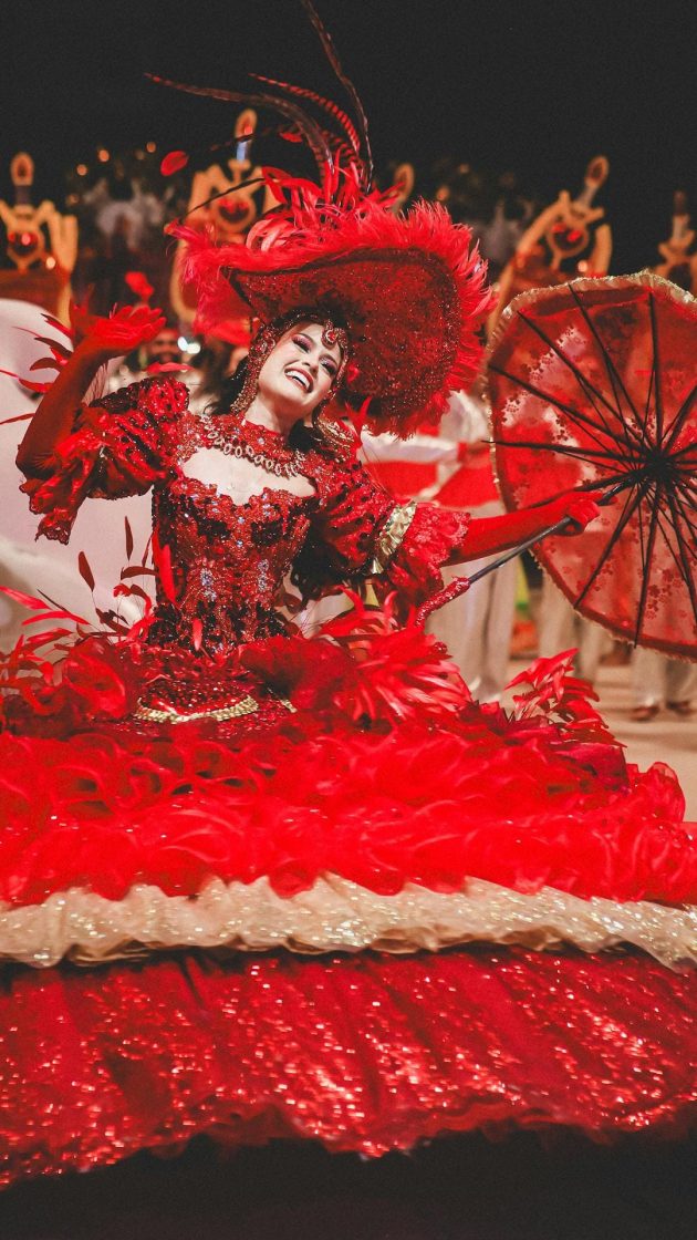 Dançarina do Boi Garantido se apresenta com vestido vermelho ornamentado, sombrinha decorada e acessórios brilhantes durante espetáculo no Bumbódromo