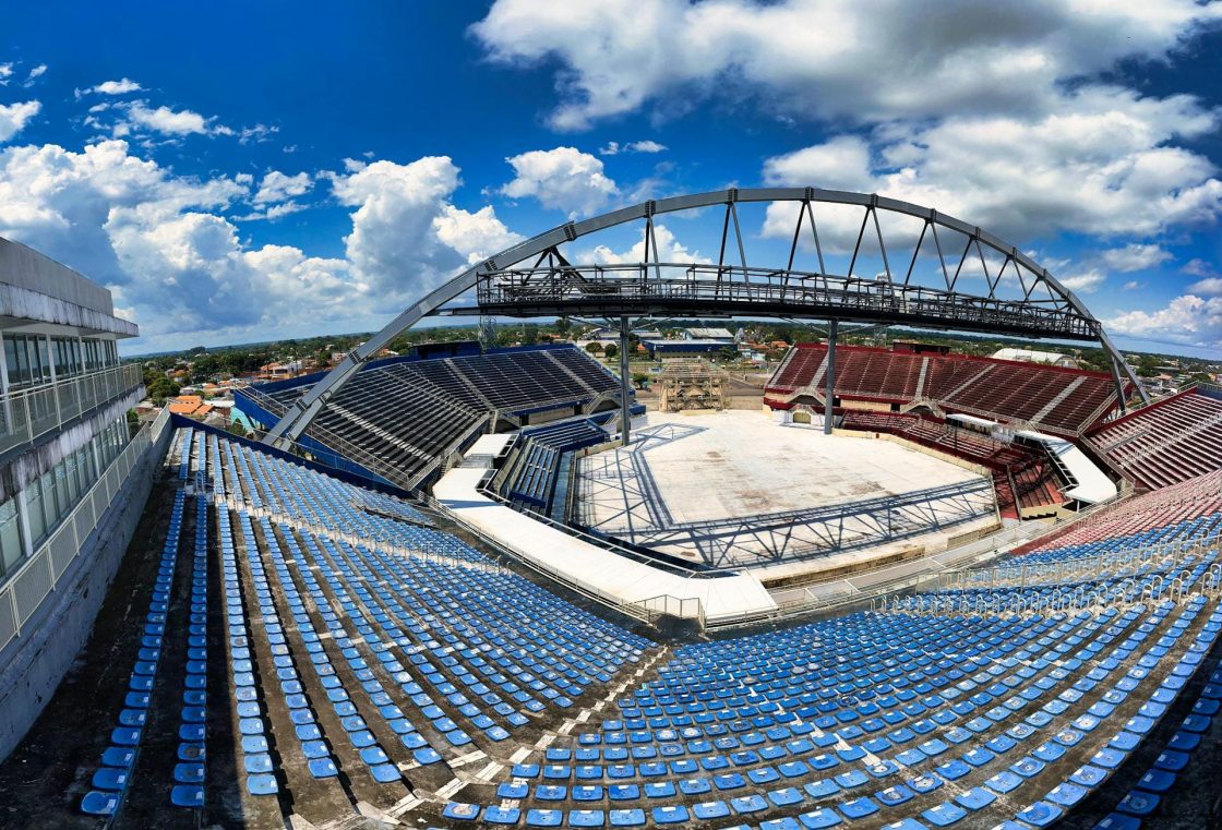 Vista panorâmica interna do Bumbódromo, com arquibancadas azuis e vermelhas divididas entre Caprichoso e Garantido, cobertura metálica arqueada e arena central vazia sob céu azul com nuvens