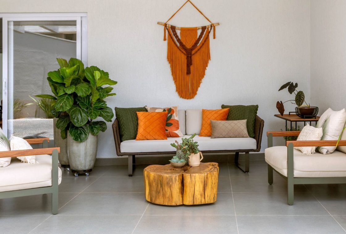 Ambiente de área externa que representa o conceito de casa com cara de casa. Ele conta com mesa de centro feita de madeira bruta, vaso de cimento com planta e macramê laranja na parede