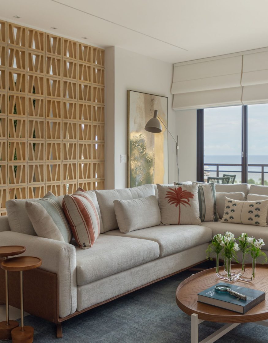Sala de estar com sofá de linho e almofadas estampadas em tons terrosos, com cobogó, quadro abstrato dourado e mesa de centro redonda em madeira com flores brancas e livro