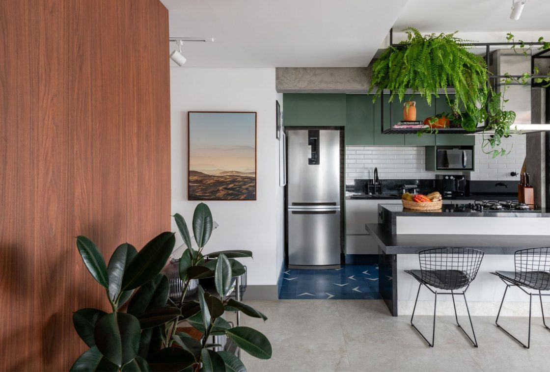 Cozinha integrada com armários verde-musgo, ladrilho hidráulico azul no piso, bancada preta e duas plantas de grande porte suspensas sobre a ilha 