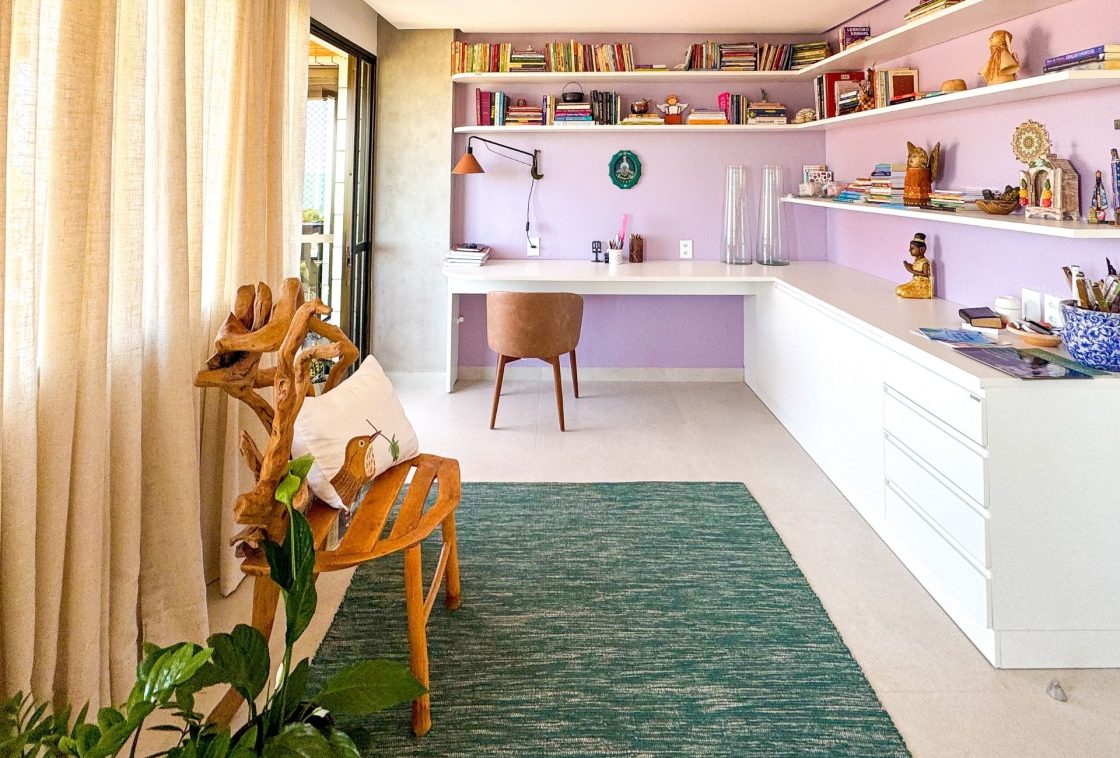 Home office com paredes lilás, prateleiras repletas de livros e objetos colecionáveis, mesa branca em "L", cadeira esculpida em madeira orgânica, tapete verde e cortina de linho bege