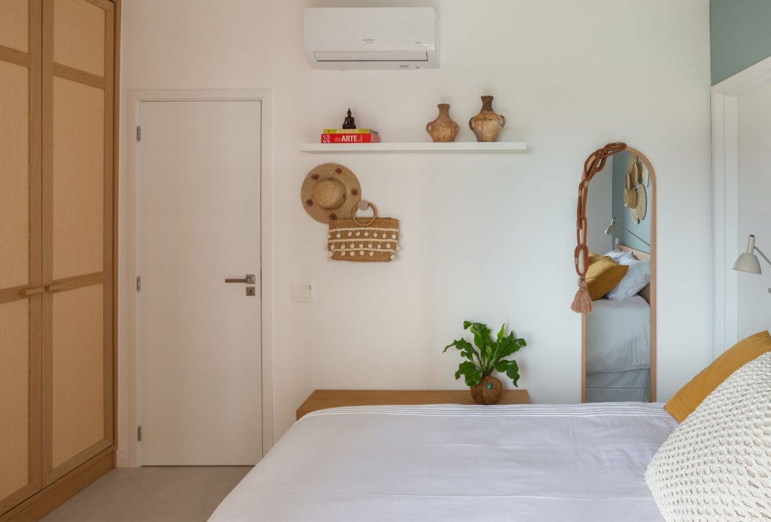 Quarto com cama de casal, prateleira com vasos de cerâmica, livro de arte, chapéu de palha e bolsa de praia pendurados na parede