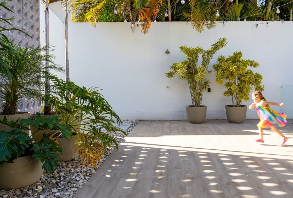 Área externa com piso de porcelanato amadeirado, vasos grandes com plantas tropicais, parede branca, cordão de luzes e uma criança correndo ao fundo com vestido colorido