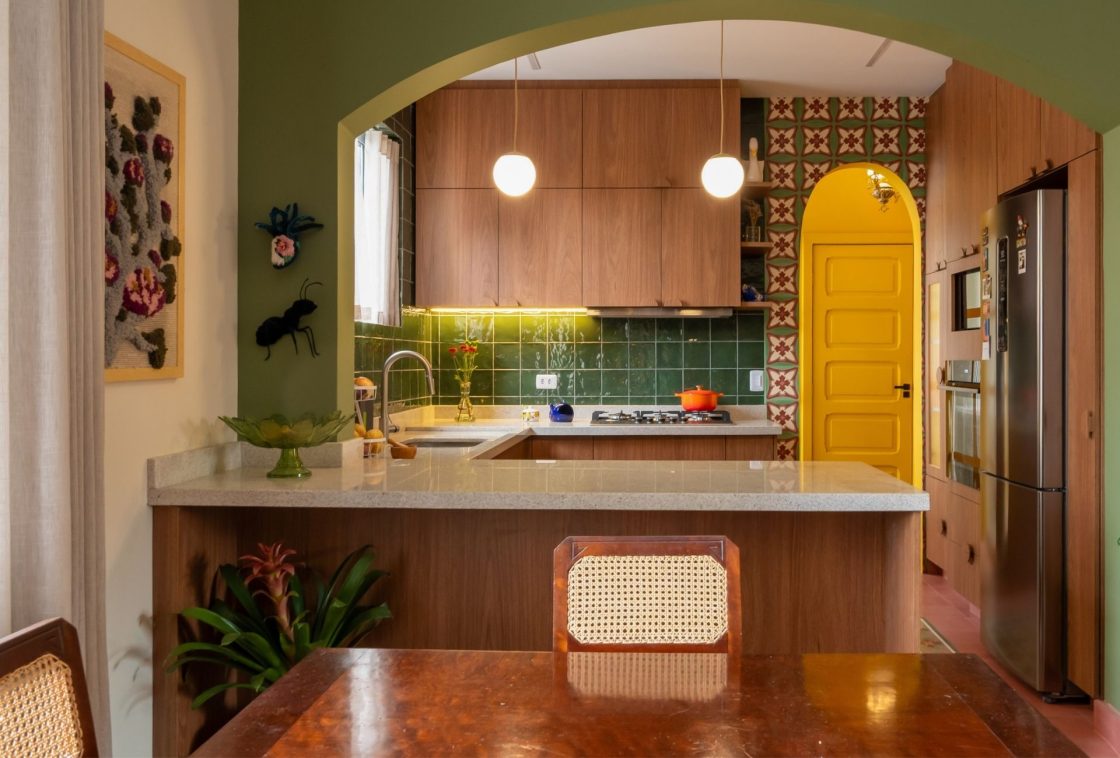 Cozinha integrada com paredes em verde-musgo, porta em arco amarela, backsplash de revestimento cerâmico verde, marcenaria em madeira escura e revestimento estampado na parede lateral