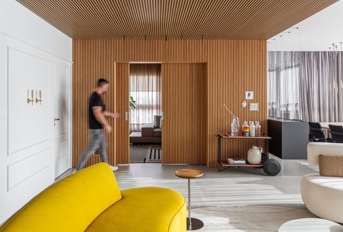 Sala de estar moderna com uma grande parede divisória revestida por painéis ripados de madeira. No primeiro plano, destaca-se a curva de um sofá em tecido de veludo amarelo vibrante