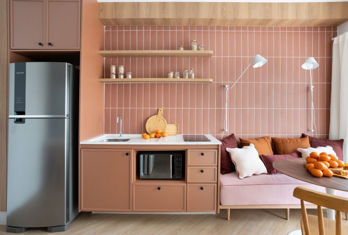 Cozinha compacta e planejada de um apartamento estúdio. A parede da bancada é revestida com revestimentos retangulares em tom rosa queimado, dispostos verticalmente com rejunte branco