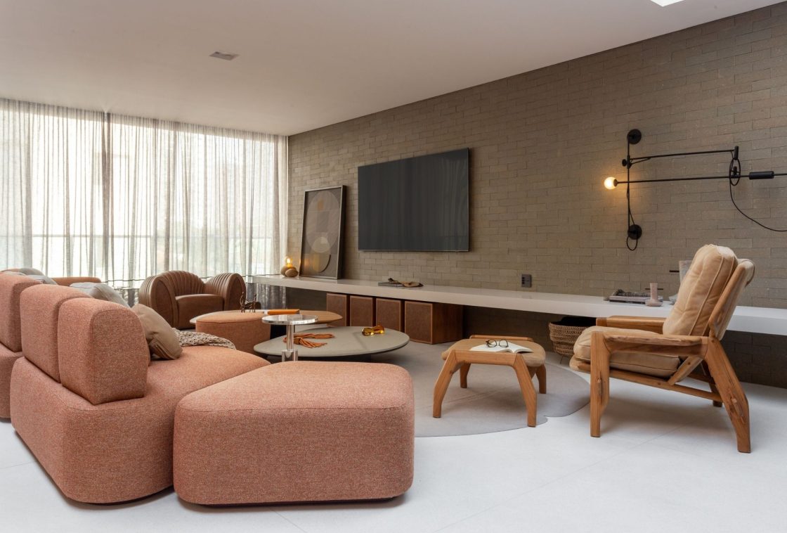 Living minimalista caloroso, com um sofá modular estofado em tom pêssego. Ao fundo, a parede principal é revestida com tijolinhos aparentes em tom marrom, onde a TV está instalada