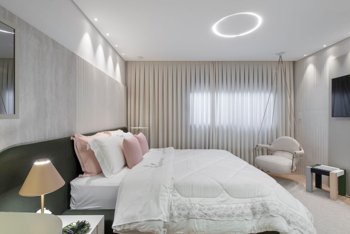 Quarto em tons neutros, com parede com cortina ao fundo e cama de casal com travesseiros rosas