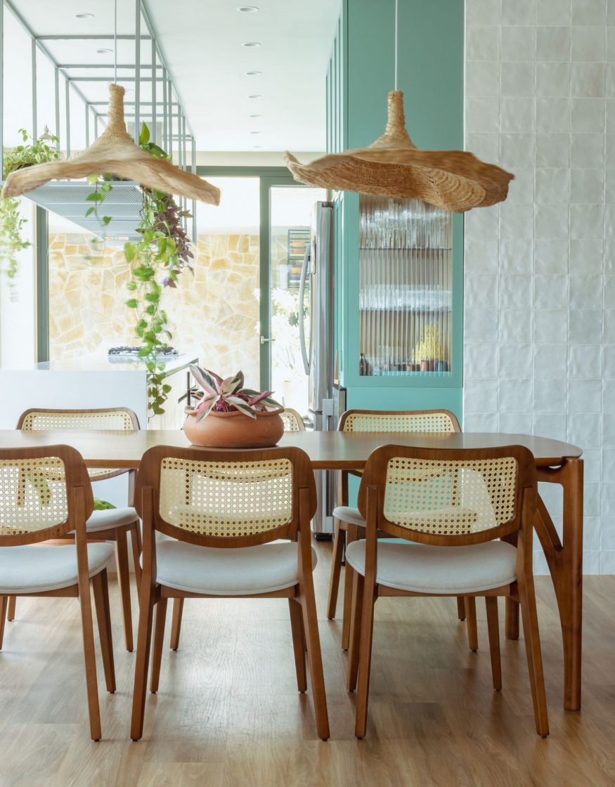 Sala de jantar integrada à cozinha com estética litorânea e suave. No centro, uma mesa de madeira clara é cercada por cadeiras com encosto de palhinha natural