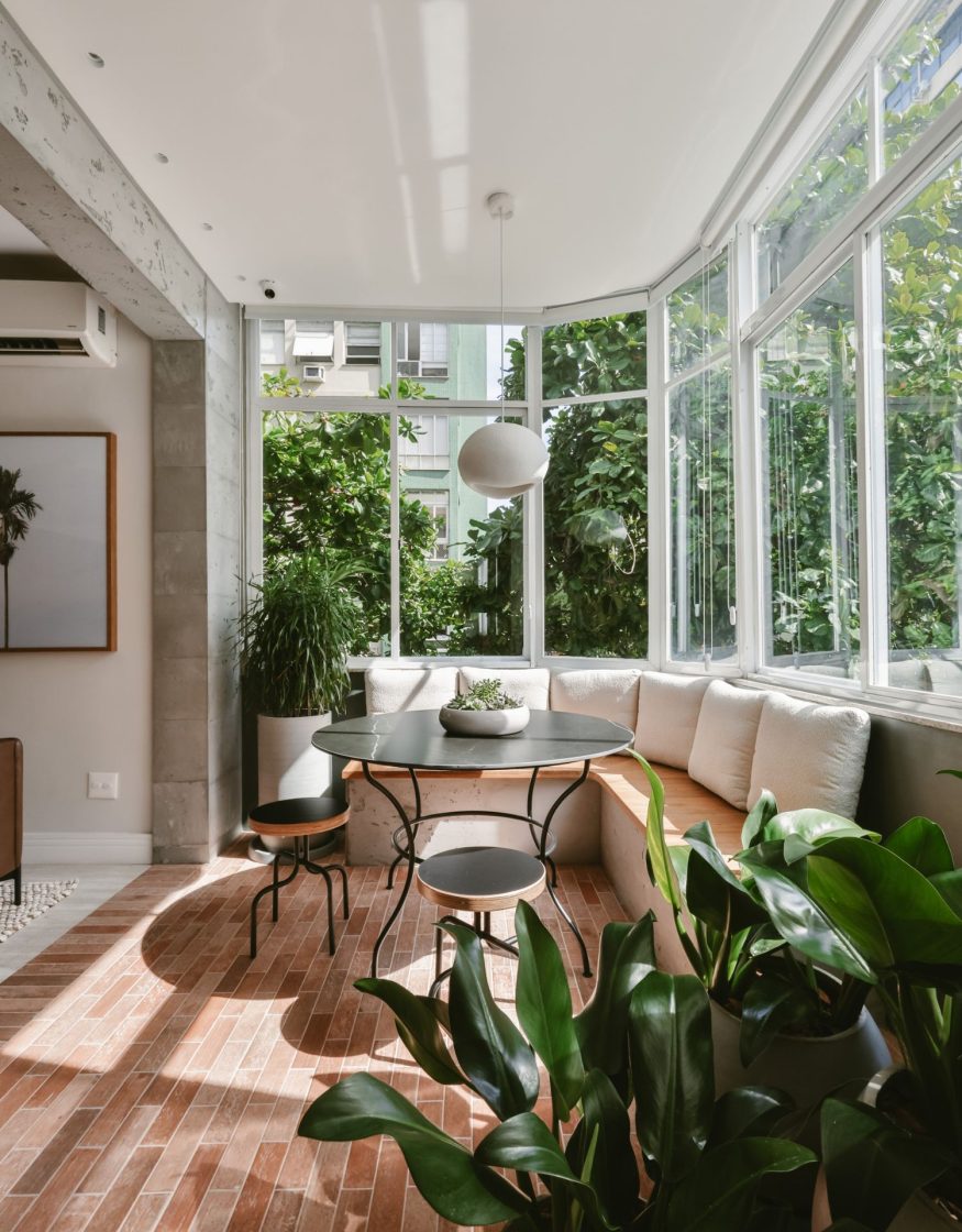 Varanda de apartamento urbano bem iluminada pela luz solar. O piso é revestido com pequenos porcelanatos em formato retangular de tom terracota, assentados em padrão de tijolinhos