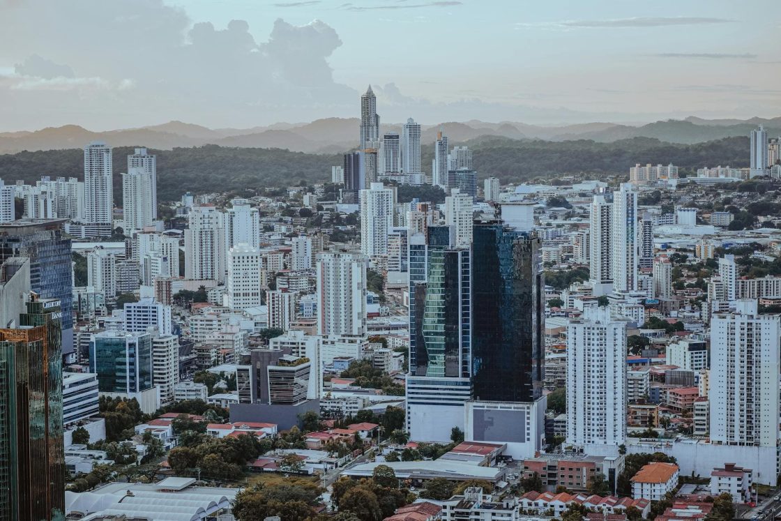 Vista panorâmica da Cidade do Panamá, com grande concentração de arranha-céus e edifícios residenciais, cercados por áreas verdes e montanhas ao fundo sob céu nublado no fim da tarde