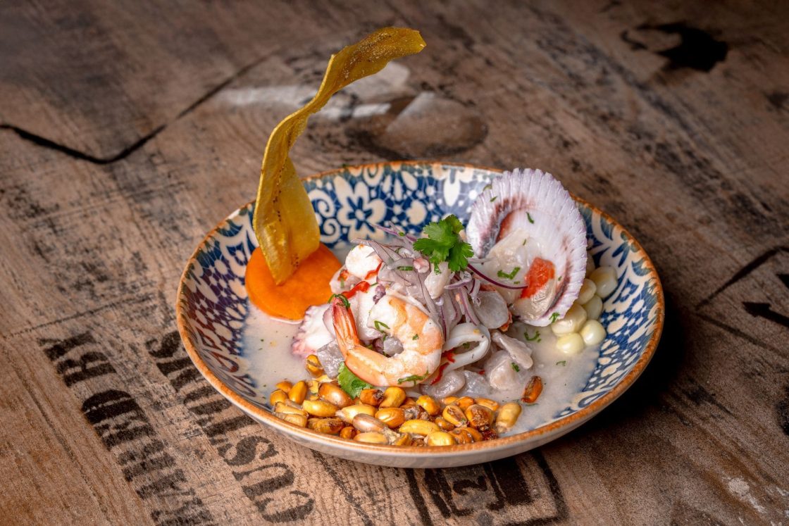Prato de ceviche panamenho servido em tigela decorada com camarão, lula, milho, cebola roxa e chips crocantes sobre mesa de madeira rústica