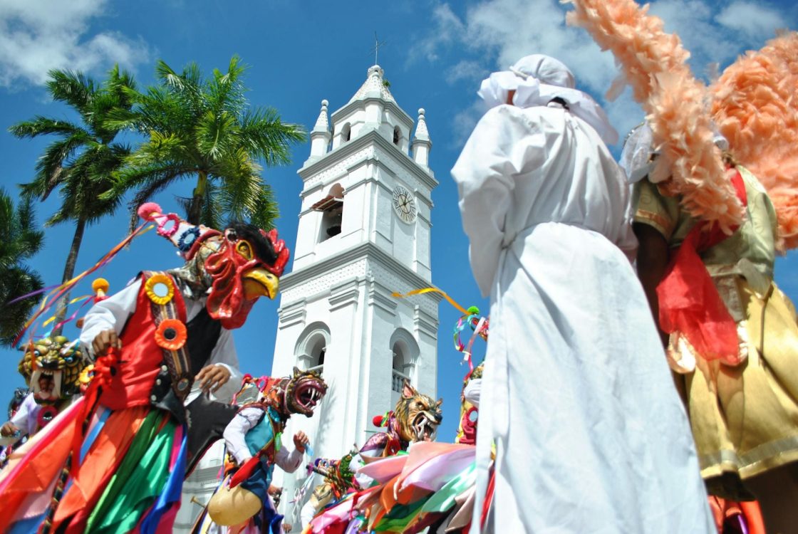Festival tradicional panamenho, com pessoas usando fantasias coloridas e máscaras diante de igreja branca, sob céu azul e palmeiras tropicais