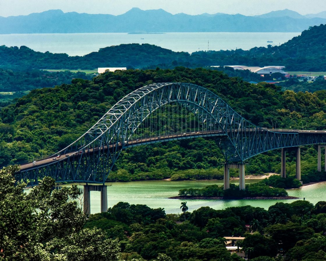 Vista aérea da Ponte das Américas, atravessando área verde e águas do Canal do Panamá, com montanhas ao fundo sob céu claro