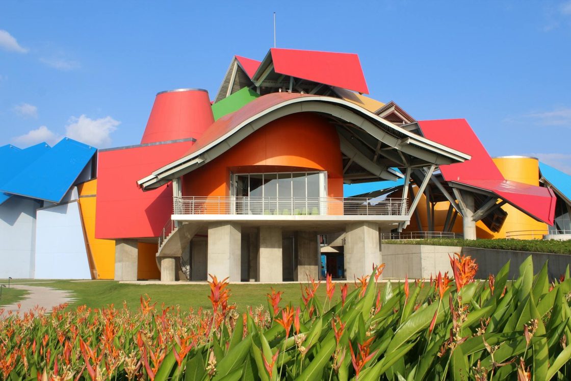 Biomuseo, projetado por Frank Gehry, com estrutura contemporânea colorida em tons de vermelho, azul, amarelo e laranja, cercada por jardim tropical sob céu azul claro