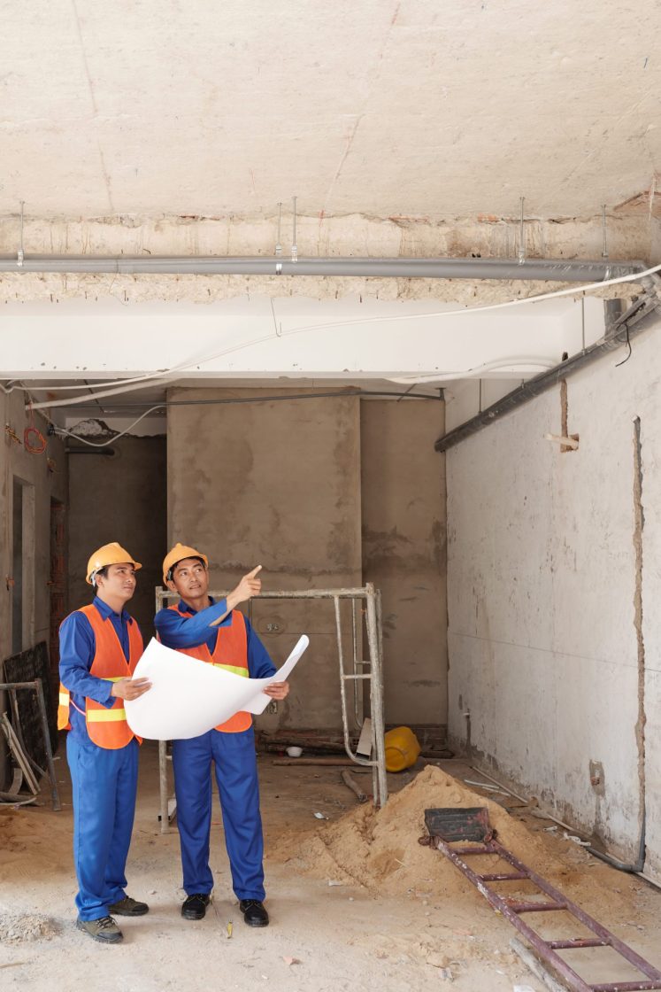Dois trabalhadores com uniforme azul, colete laranja e capacete, ambos em obra na fase de embosso