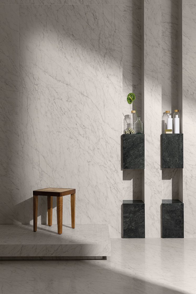 Ambiente todo revestido em porcelanato que reproduz mármore branco, com luz natural entrando pelo lado direito, dois balcões em porcelanato verde-escuro ao fundo e um banco de madeira mais à frente