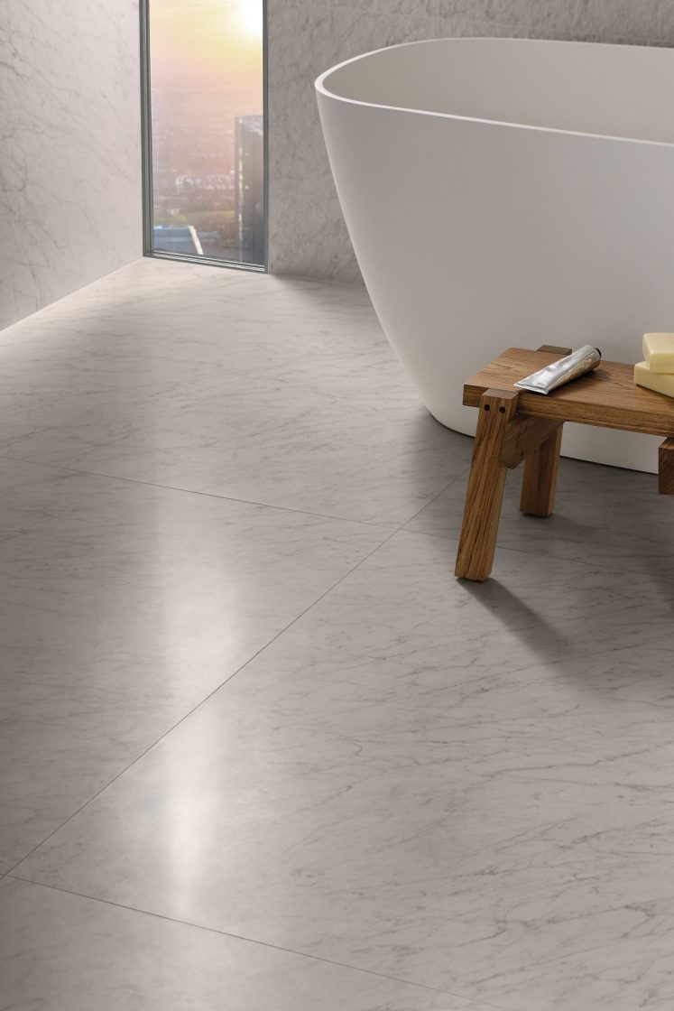 Detalhe de ambiente todo revestido em porcelanato que reproduz mármore branco, com ponta de banheira de imersão aparecendo à direita, ao lado de janela de vidro estreita à esquerda
