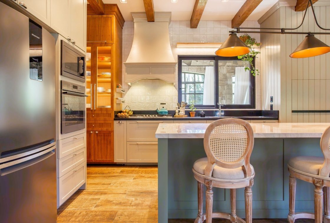 Torre quente em marcenaria de madeira com forno e micro-ondas embutidos em cozinha estilo farmhouse, com vigas aparentes e ilha central
