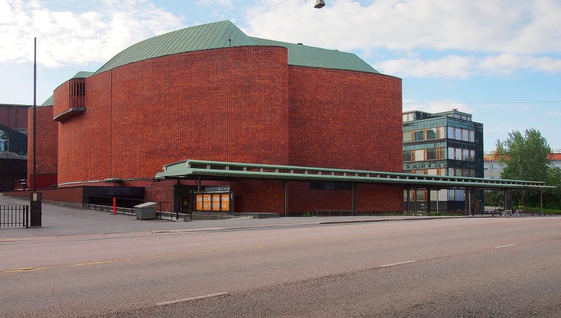 Vista externa do centro cultural projetado por Alvar Aalto em Helsinque, com volumes curvos em tijolos vermelhos, cobertura metálica esverdeada e marquise voltada para a avenida