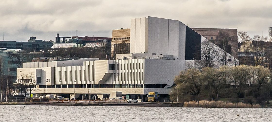 Vista do Finlandia Hall às margens da baía de Töölö, em Helsinque, mostrando o edifício revestido em mármore branco projetado por Alvar Aalto em meio à paisagem urbana e arborizada da capital finlandesa