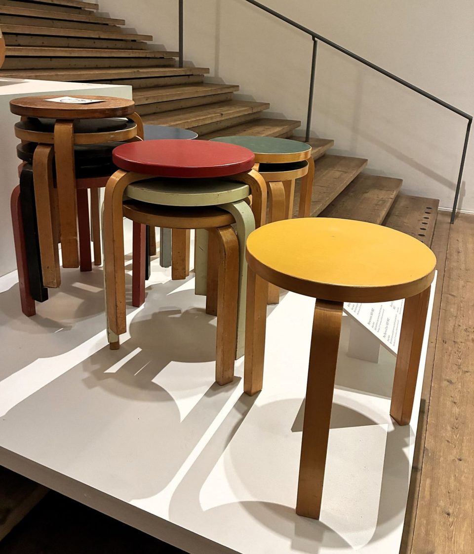 Conjunto de banquetas Modelo 60 desenhadas por Alvar Aalto, com assentos circulares coloridos e pernas curvas em madeira laminada, expostas em um ambiente interno com escada de madeira ao fundo