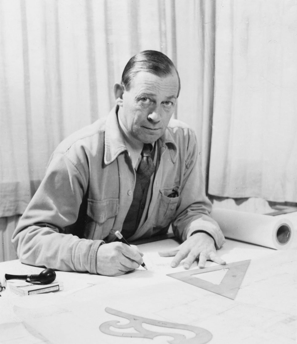Fotografia em preto e branco de Alvar Aalto sentado à mesa de trabalho, desenhando com lápis sobre uma prancheta cercada por instrumentos de desenho arquitetônico
