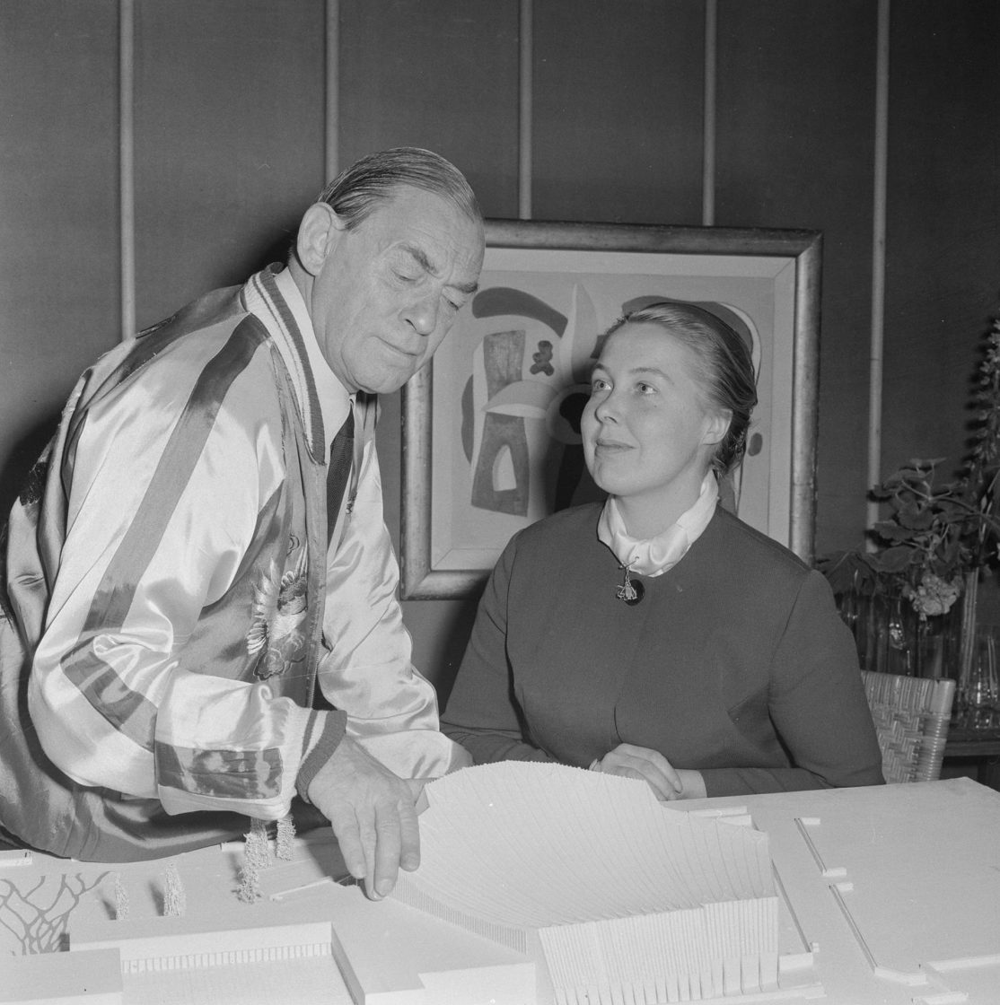 Fotografia em preto e branco de Alvar Aalto e da arquiteta Elissa Mäkiniemi observando uma maquete arquitetônica sobre uma mesa em um ambiente interno com quadro decorativo ao fundo