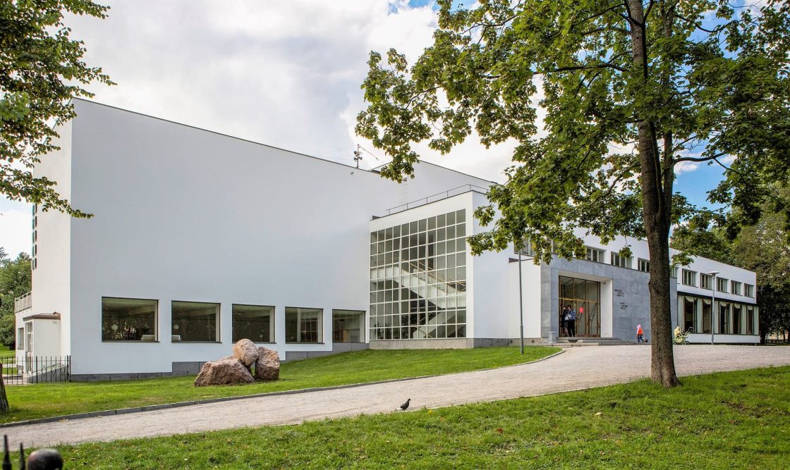 Vista externa da Biblioteca de Viipuri, edifício branco de linhas modernas projetado por Alvar Aalto, cercado por árvores e gramado em dia parcialmente nublado