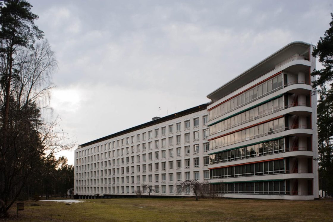 Vista externa do Sanatório de Paimio, edifício modernista branco projetado por Alvar Aalto, com alas alongadas, varandas curvas e área gramada cercada por árvores altas