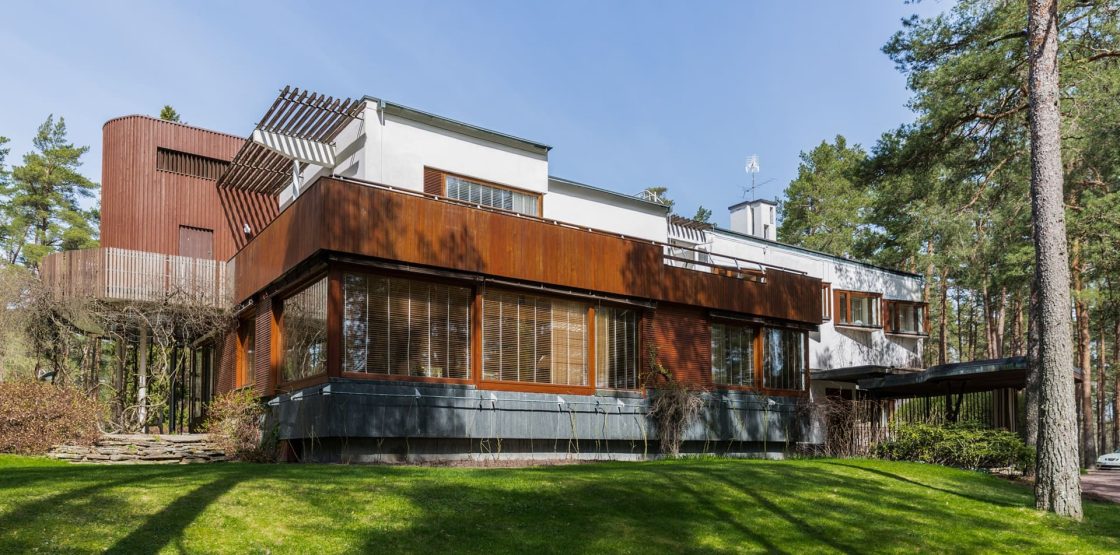 Vista externa da Villa Mairea, residência projetada por Alvar Aalto em meio a uma área arborizada, com volumes brancos, revestimentos em madeira e amplas superfícies envidraçadas