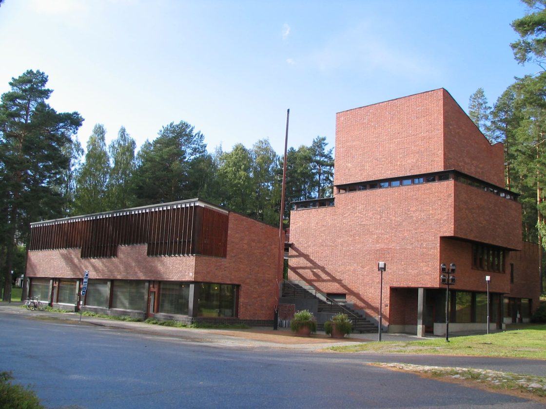 Vista externa da Prefeitura de Säynätsalo, projetada por Alvar Aalto, com volumes em tijolos aparentes, escadaria externa e bosque de pinheiros ao redor do edifício