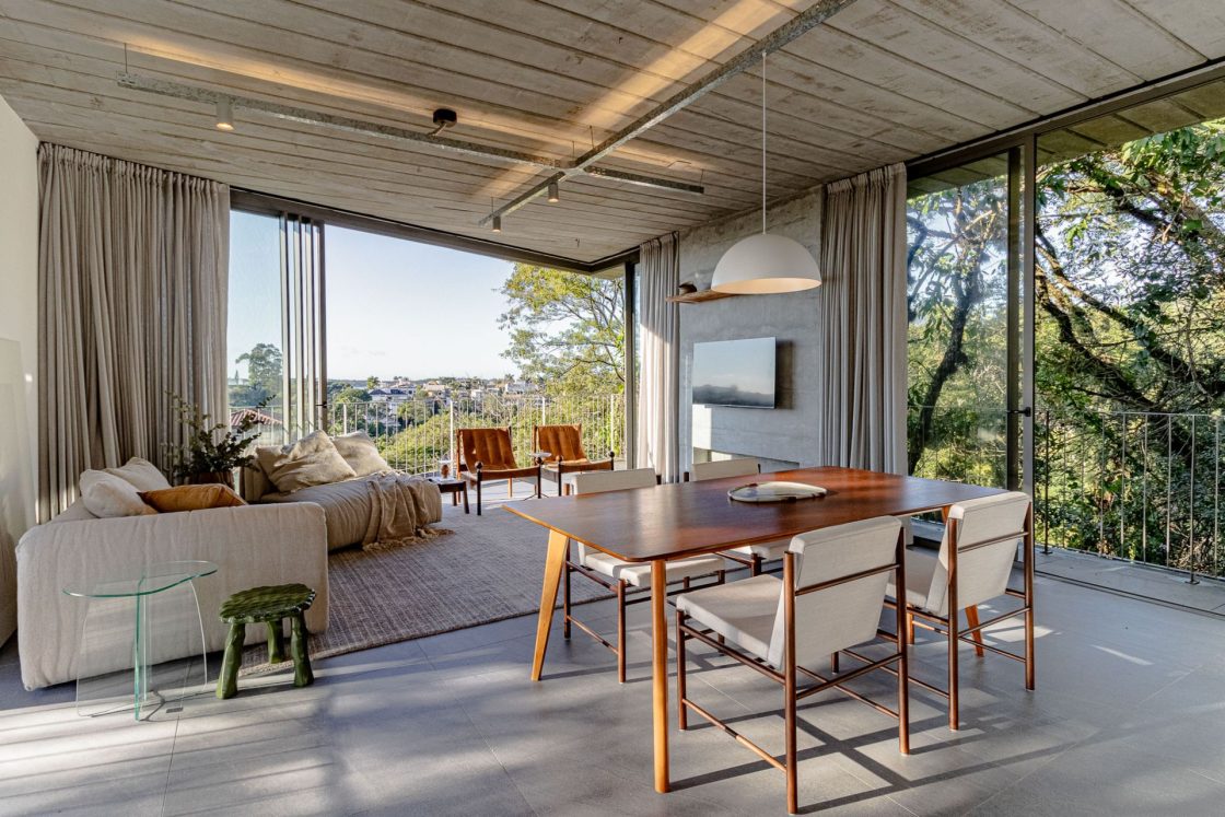 Living com televisão e mesa de jantar de madeira com quatro cadeiras e estofado off white em casa com piso que reproduz pedra e tem vista para área verde