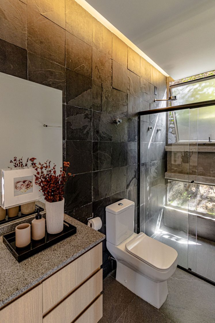 Banheiro com piso que reproduz limestone em tom de cinza, bancada cinza e parede em revestimento que reproduz pedra em tom escuro