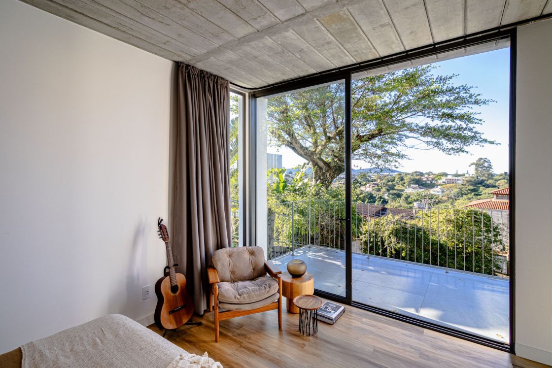 Quarto com piso amadeirado e varanda com piso que reproduz pedra cinza, com grande pano de vidro integrando ambientes