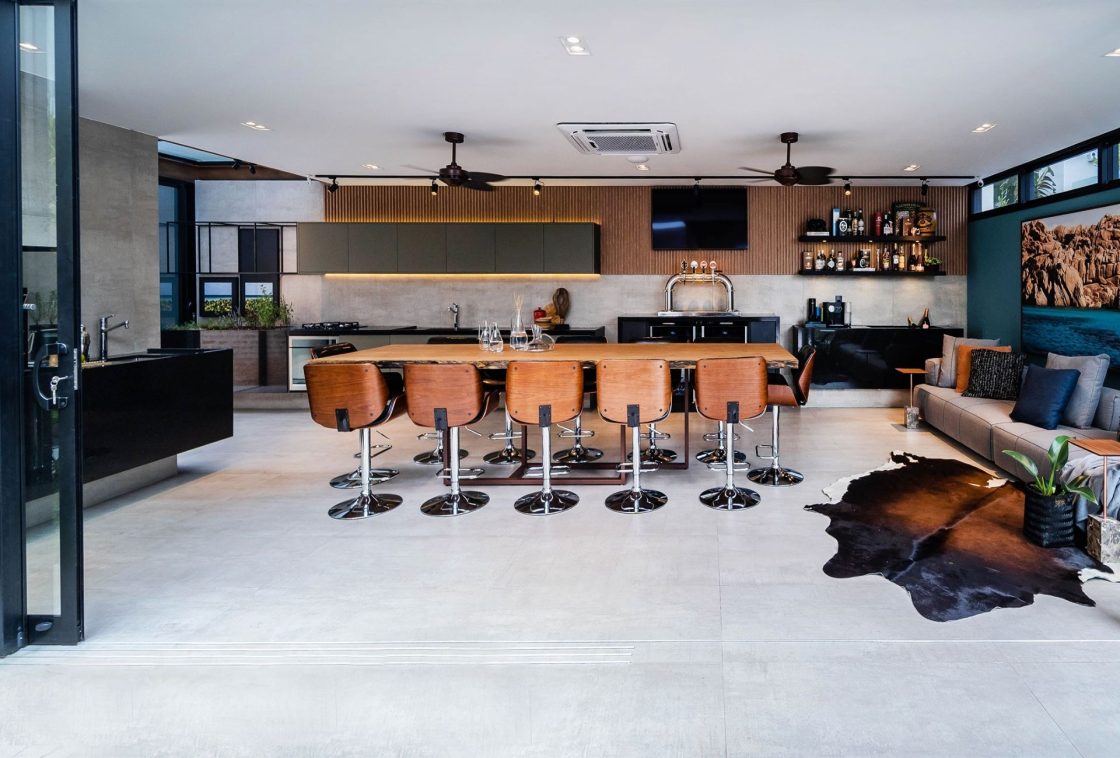 Ambiente de lazer amplo e integrado, que une sala de estar e área gourmet de forma contínua. No centro do espaço, há uma grande mesa retangular de madeira maciça, acompanhada por diversas banquetas giratórias com encosto de madeira