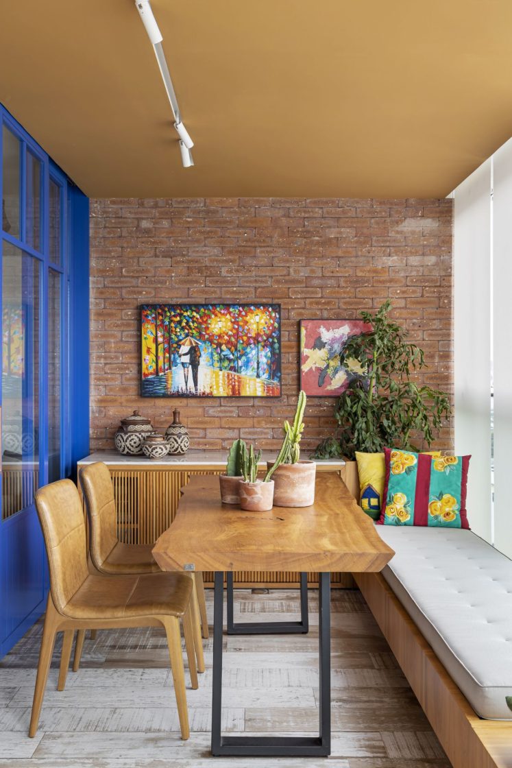 Sala de jantar com mesa de madeira, banco de um lado e cadeiras em couro marrom do outro, com parede de tijolinhos decorada com quadros ao fundo
