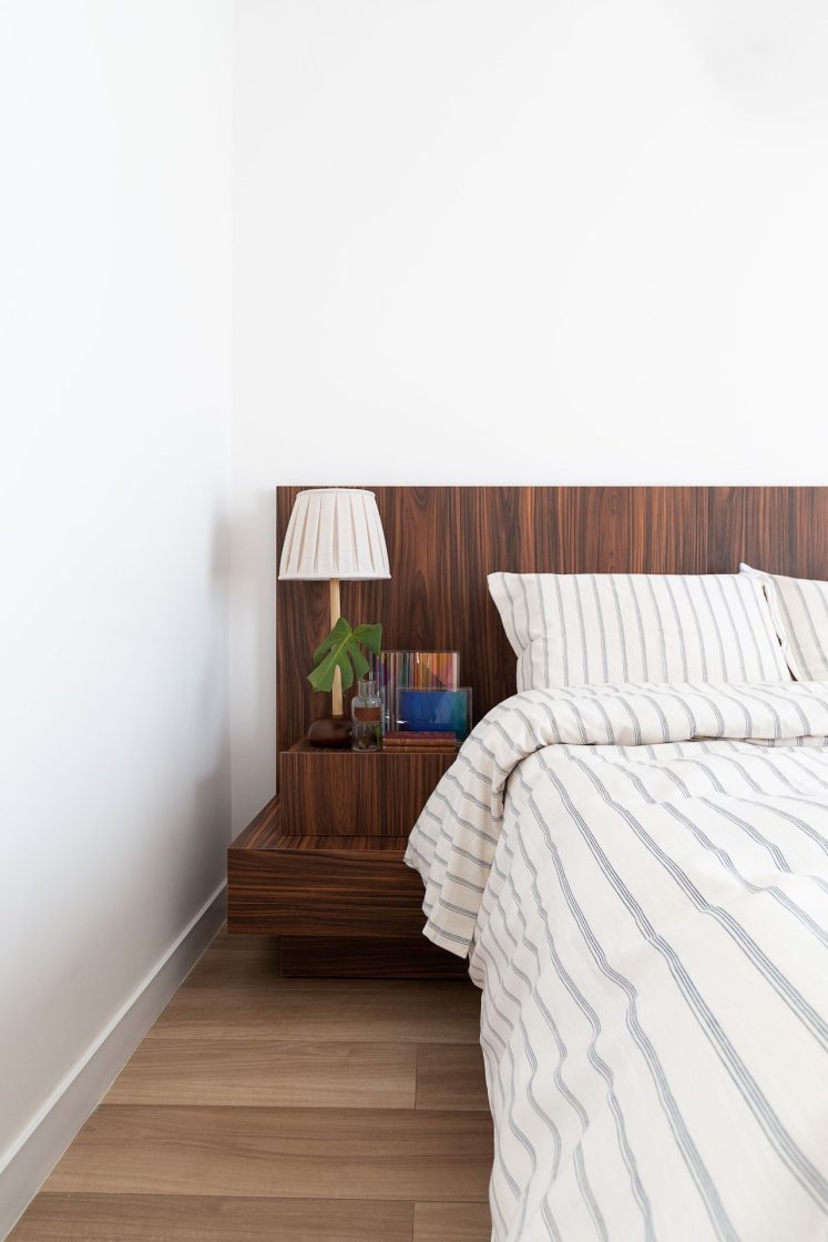 Detalhe de cama de casal com cabeceira e mesa lateral amadeirada, piso que reproduz madeira e rodapé branco