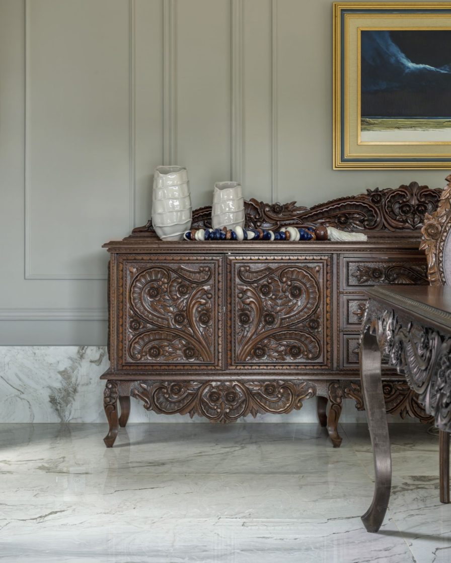 Detalhe de buffet antigo em madeira escura, todo trabalhado, em ambiente com piso que reproduz mármore branco