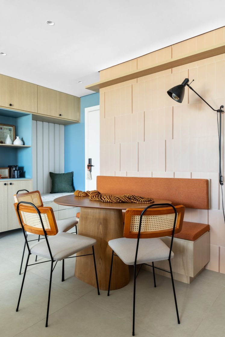 Cozinha com mesa de jantar oval com banco e três cadeiras, em frente a parede com revestimento bege com filetes que sobressaem