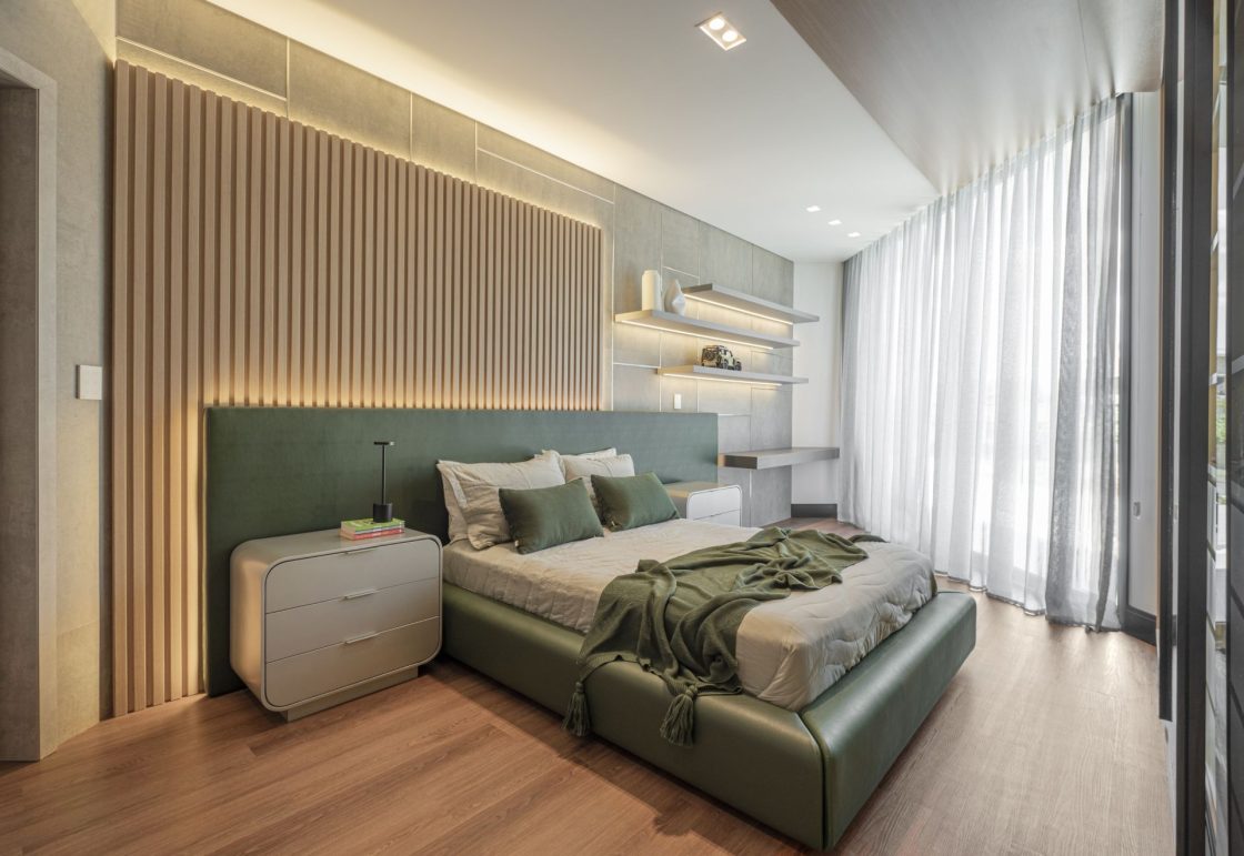 Quarto com piso amadeirado e detalhes em verde-escuro, como a cabeceira, que está em frente a um painel ripado