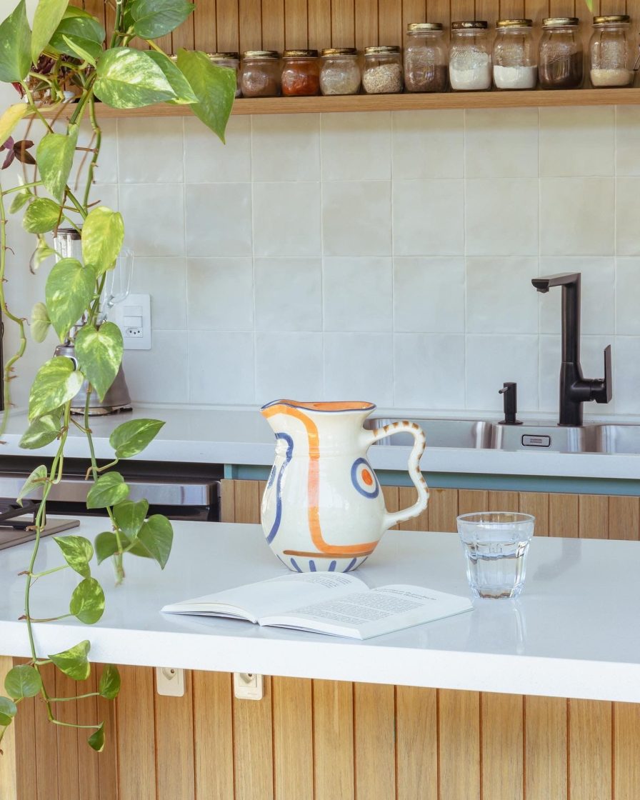 Jarra artesanal com desenhos em azul e laranja sobre bancada de ilha de cozinha com mobiliário em madeira ripada e balcão branco