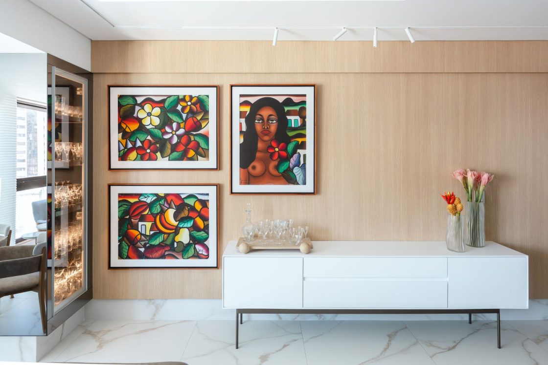 Ambiente residencial com buffet branco em frente a painel de madeira com três quadros em tons predominantes de verde e vermelho