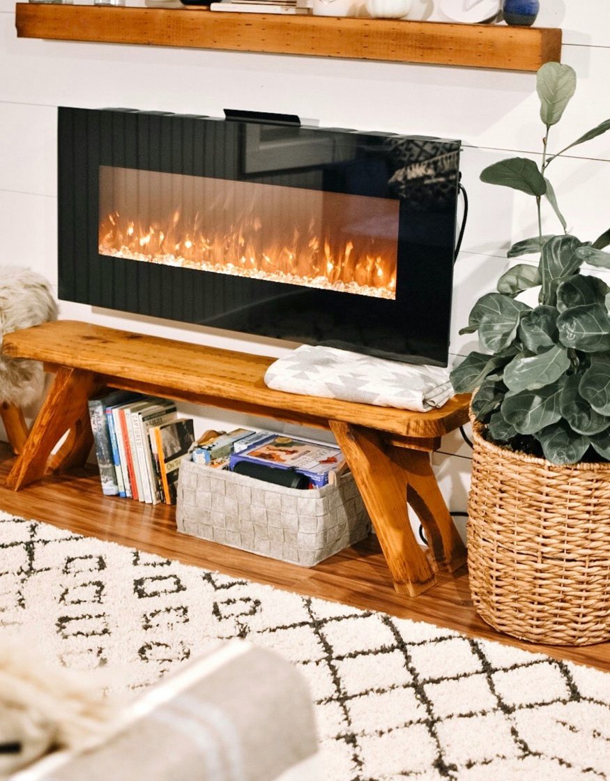Ambiente aconchegante e despojado, com um banco em frente a uma lareira elétrica. Na decoração, tapete felpudo com estampa em grafismo e cachepot feito de vime