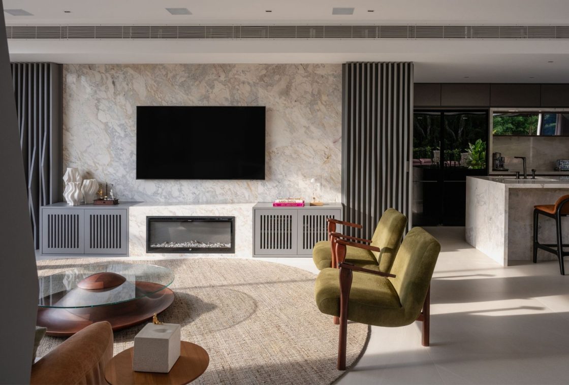 Sala de estar com lareira embutida na marcenaria embaixo da TV. Há ainda duas poltronas em tom verde-musgo, mesa de centro redonda e objetos de decoração sobre o rack