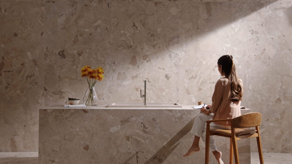 Cozinha toda em revestimento que reproduz pedra, com ilha com cuba de embutir e monocomando prateado, e mulher sentada em banqueta, de costas enquanto faz anotação em caderno